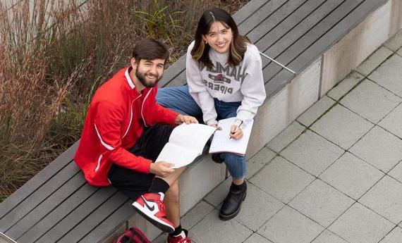 Two Hamline students sitting on a bench outside looking up at the camera smiling