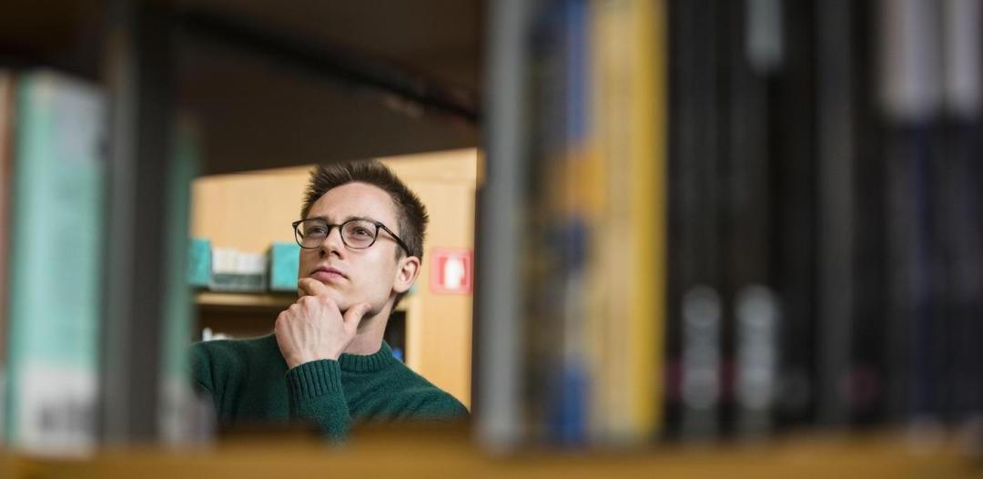 An individual wearing glasses and peering at a library shelf with their hand on their chin